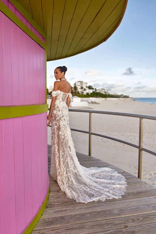 Chloe lined dress on a model near a pink beach house, showcasing an off-the-shoulder design and a flowing lace train.
