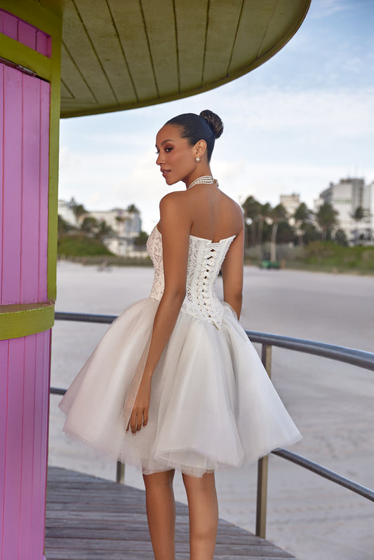 Model in a short white dress showcasing modern bridal style dresses from the back at a beachside location.