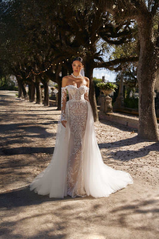 Luxurious fitted wedding dress with a detachable overskirt, showcased in an outdoor setting with trees.