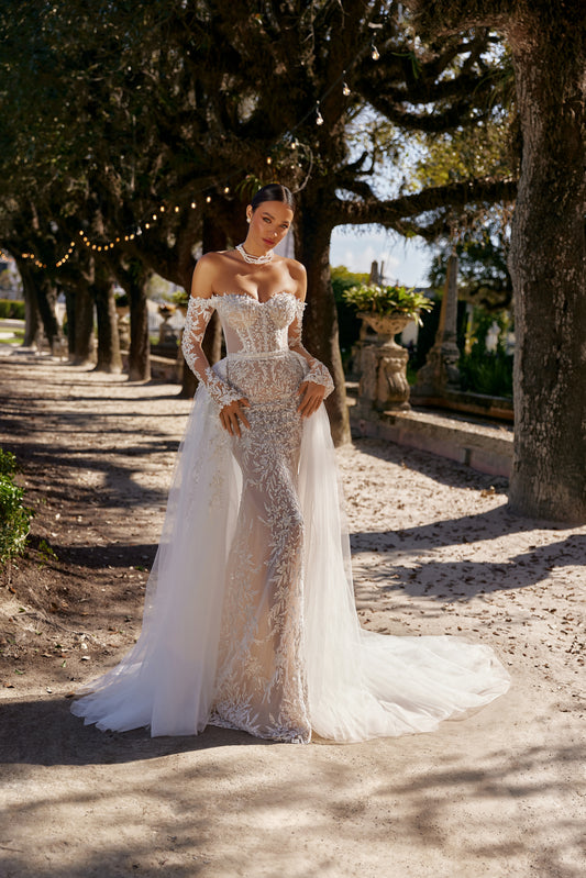 Luxurious fitted wedding dress displayed outdoors with an elegant overskirt, highlighting intricate lace details and a stunning silhouette.