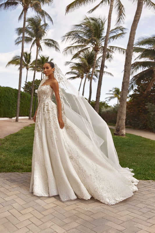 A model wearing a royal dress with cape, standing elegantly among palm trees in a picturesque outdoor setting.