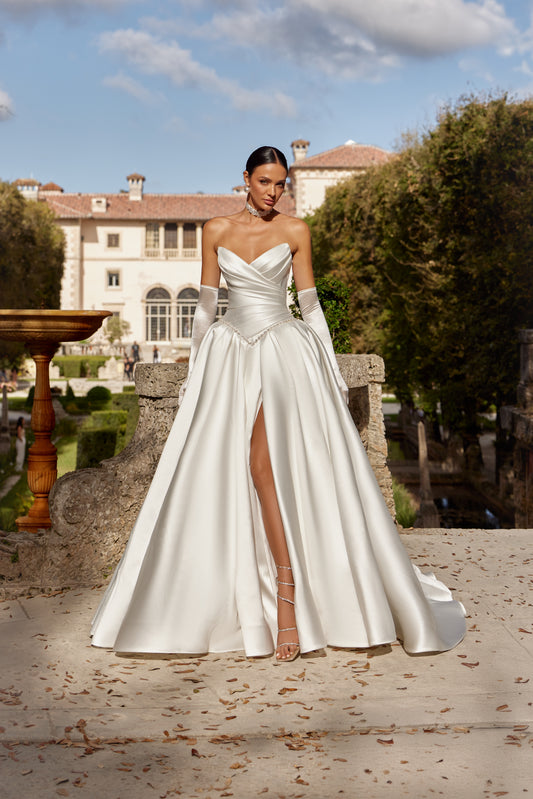 Model wearing a luxurious ball gown with a high slit, corset bodice, and elegant train, in a beautiful outdoor setting.