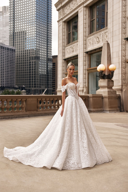 Model in a romantic A-line lace dress with an off-shoulder neckline and voluminous skirt in an urban setting.