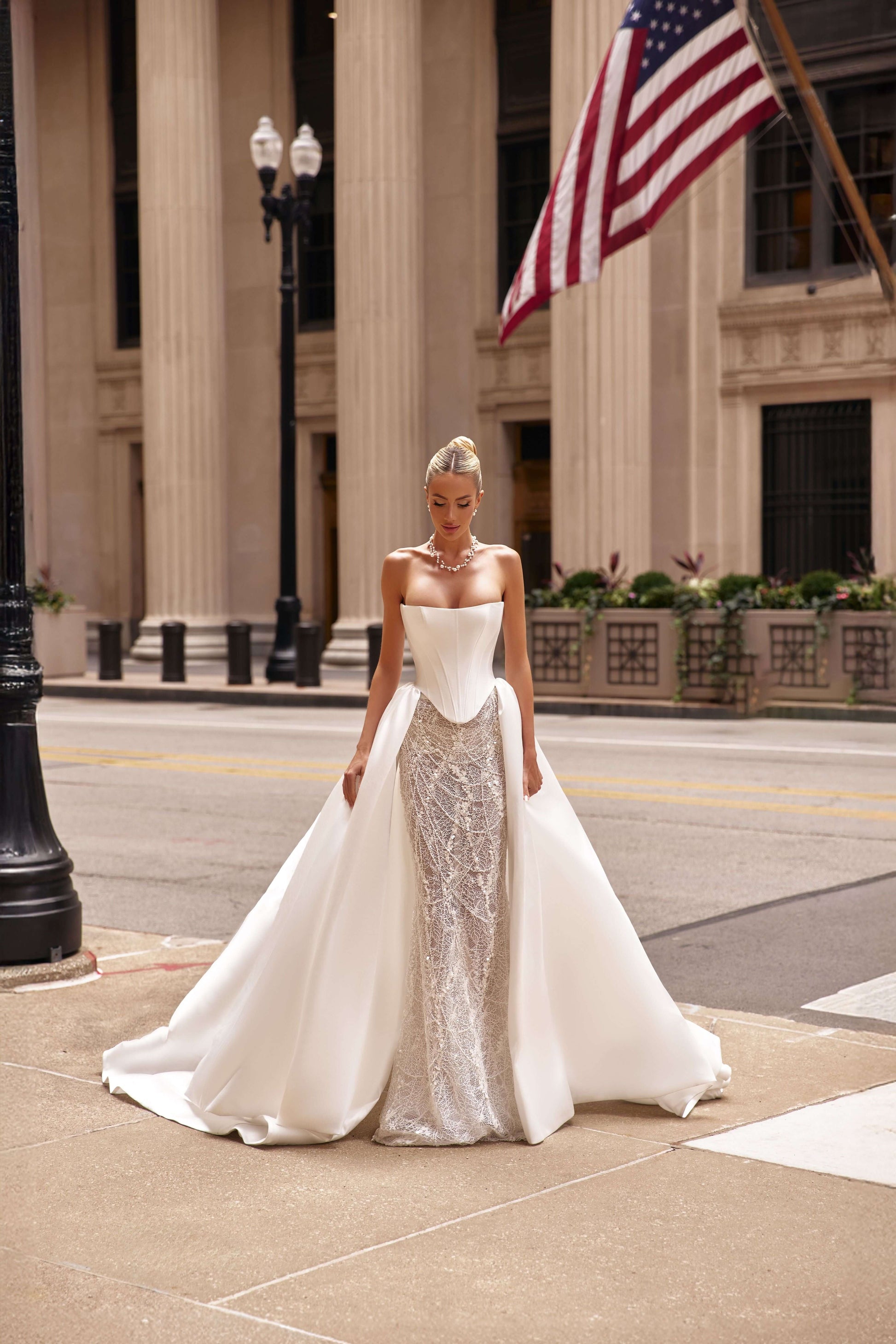Model wearing a shantung satin corset dress with an attached train, posing on a city street with an American flag in the background.