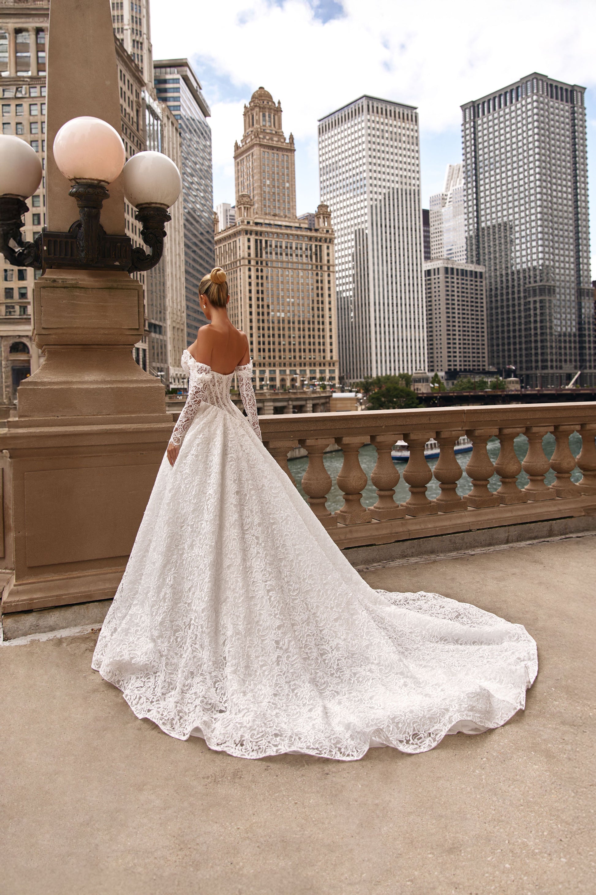Back view of a bride in a romantic A-line lace dress with a long train, standing against an urban skyline.