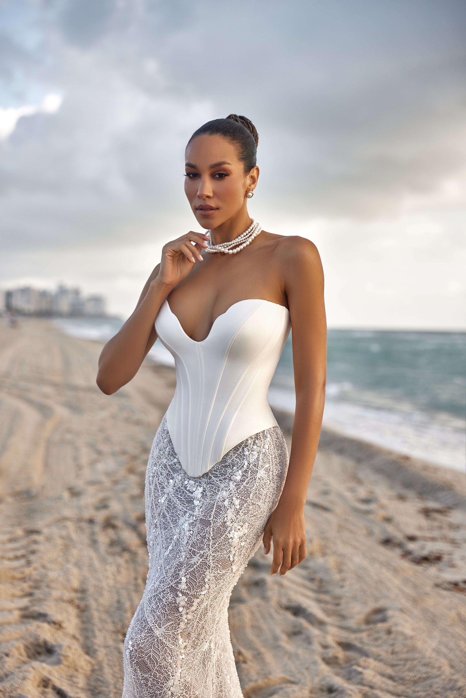Model showcasing a modern corset skirt set on a beach, featuring a strapless corset and lace skirt.