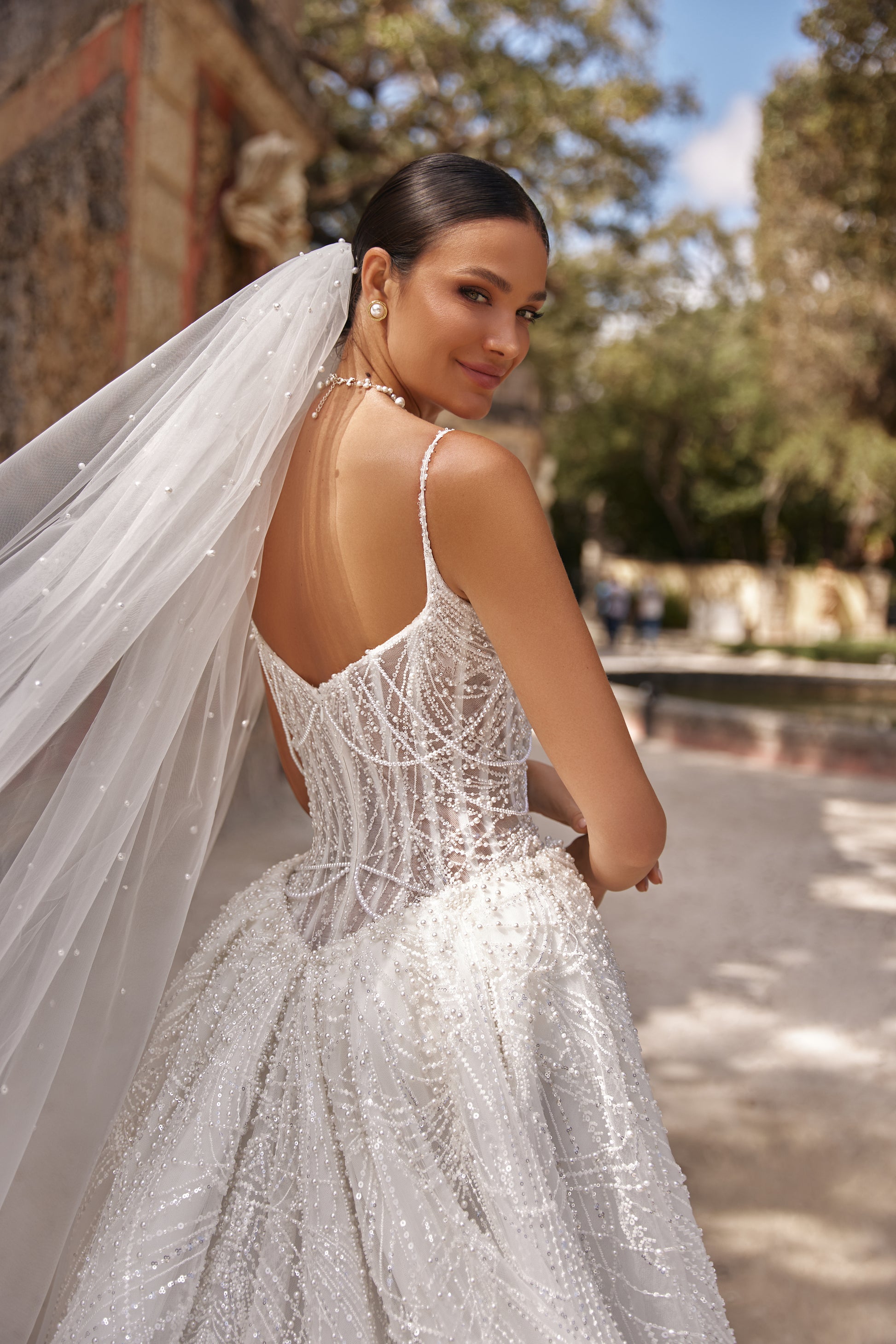 Elegant bride in a backless gown with detachable lace overskirt and long veil, showcasing intricate beading and embroidery.