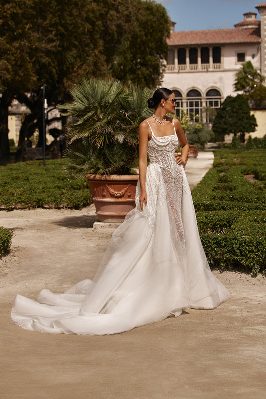 Beatrice oversized dress with detachable lace overskirt showcased in a lush garden setting with elegant architecture.