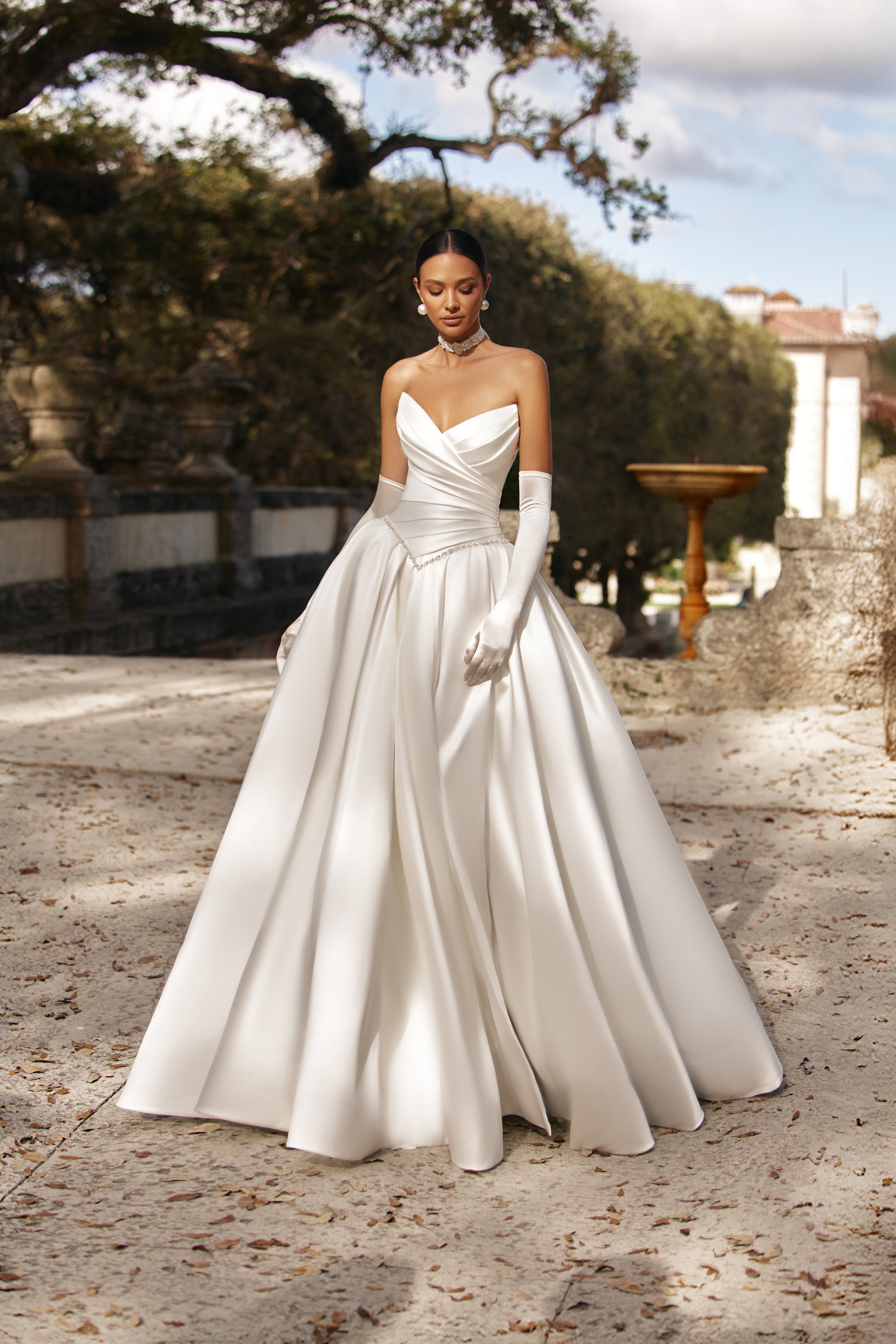 Elegant model wearing a luxurious ball gown with a corset bodice and long gloves in a picturesque outdoor setting.