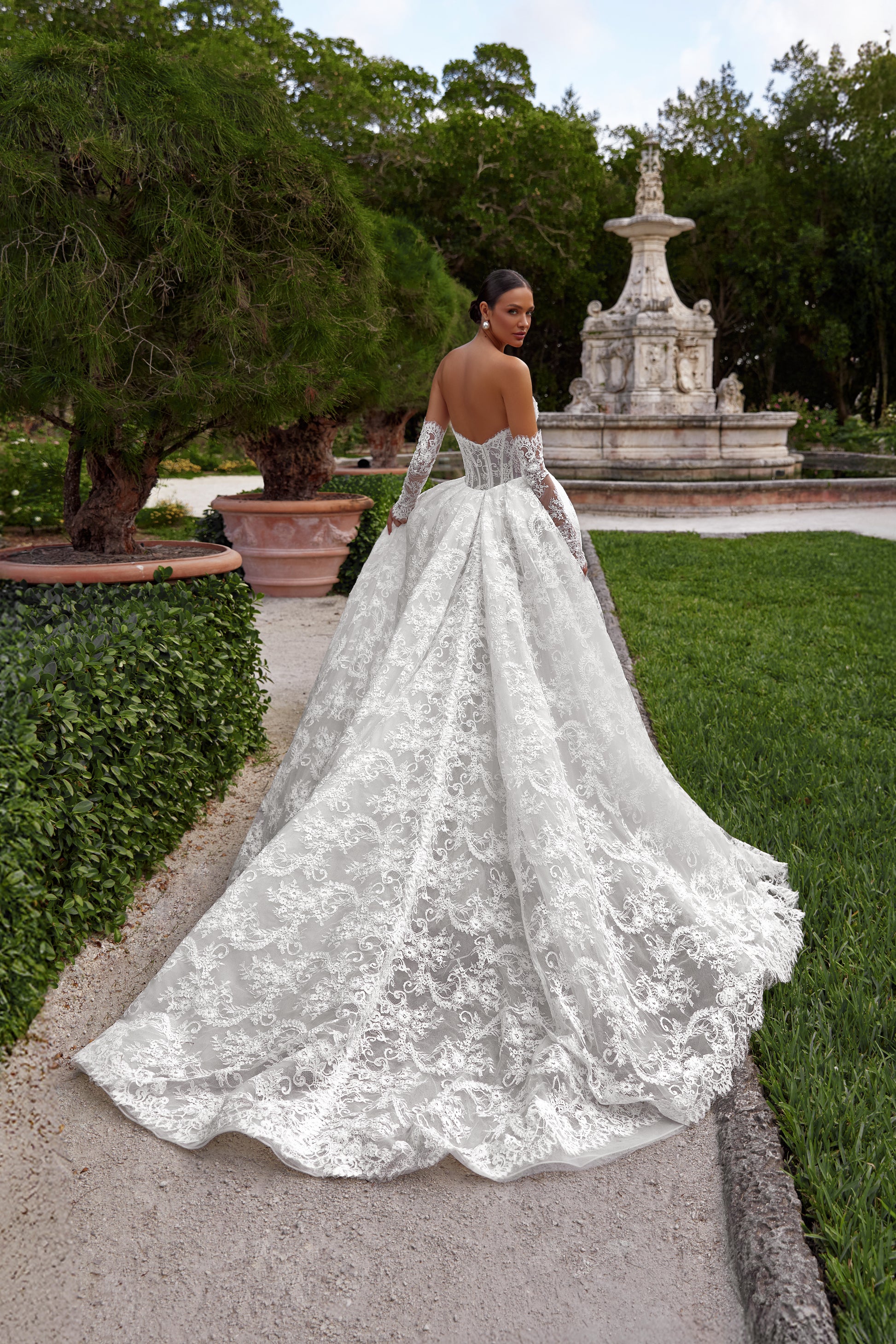 Bella wedding dress with detachable train showcased in a garden setting, featuring an elegant lace design and dramatic back.